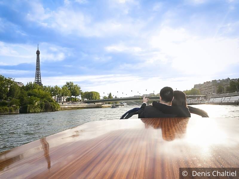 Un couple est assis à l'avant d'un bateau-mouche et tourne le dos à l'objectif. Ils regardent et pointent du doigt la tour Eiffel.